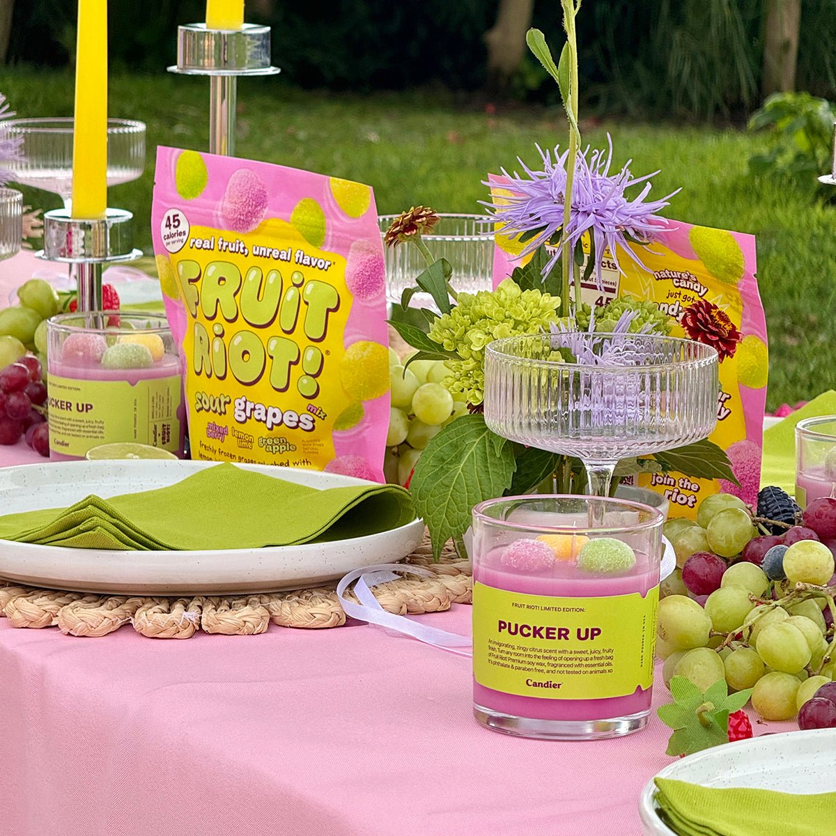 Candier Fruit Riot Sour Grapes candle styled on picnic table with citrus fruits and candycore packaging. A lux candle that blends cute pink aesthetic with food-scented dessert candle inspiration.