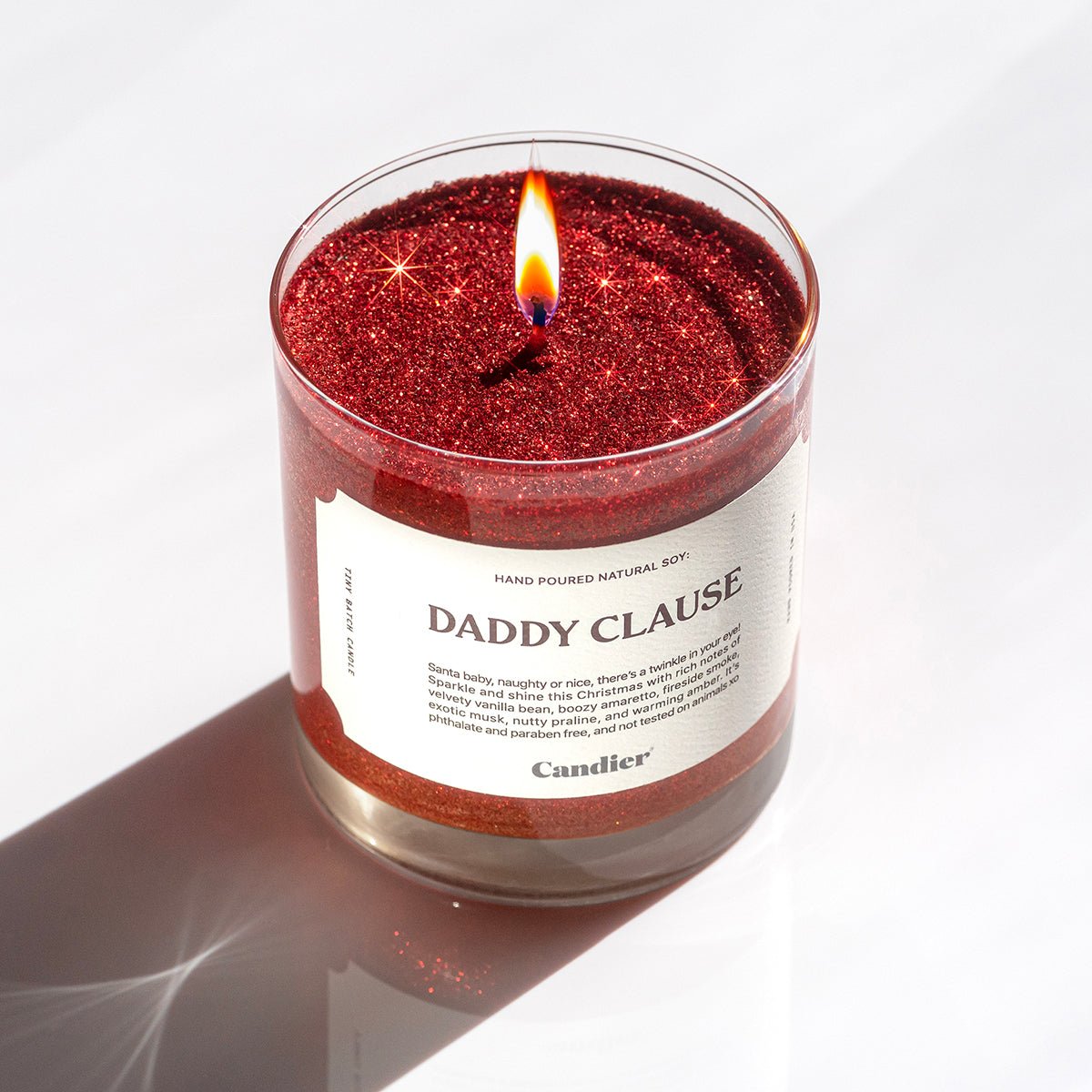 A lit festive Christmas red scented candle with plant based sparkly glitter and a label that reads Daddy Clause by Candier