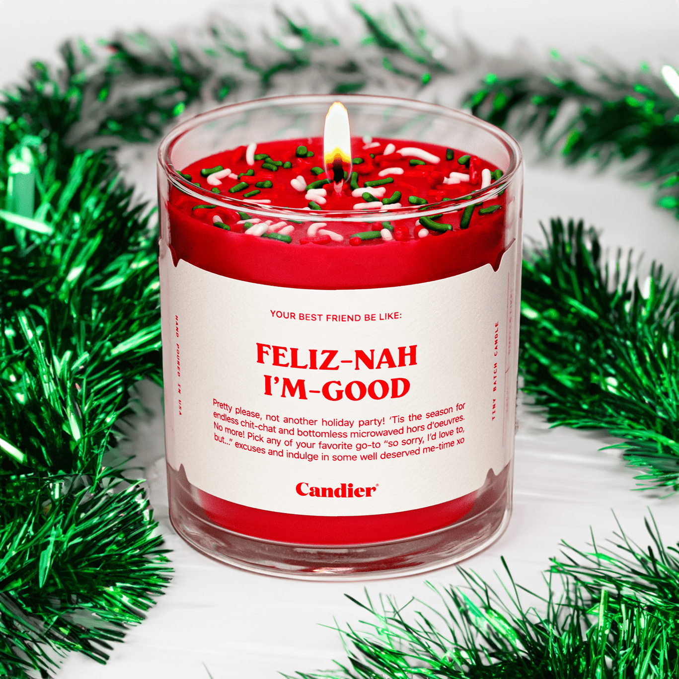 A lit Candier candle in a clear glass vessel, filled with Santa red wax, topped with red, green and white sprinkles, and a white label that reads “Feliz Nah I’m Good”. Green Christmas tinsel in the background