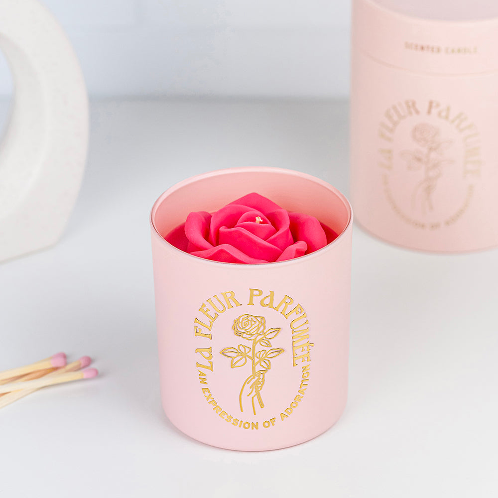 A fancy  expensive looking light pink candle with a gold emblem that reads La Fleur Parfumée An Expression of Adoration, and is topped with a hot pink sculpted wax rose