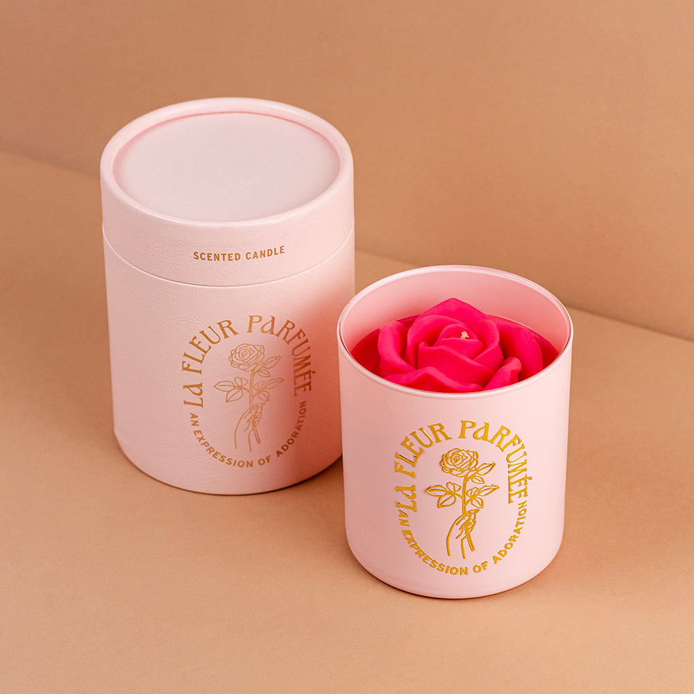 An elegant luxurious light pink candle with a gold emblem that reads La Fleur Parfumée An Expression of Adoration, and is topped with a hot pink sculpted wax rose. It sits beside an elegant luxe gift box
