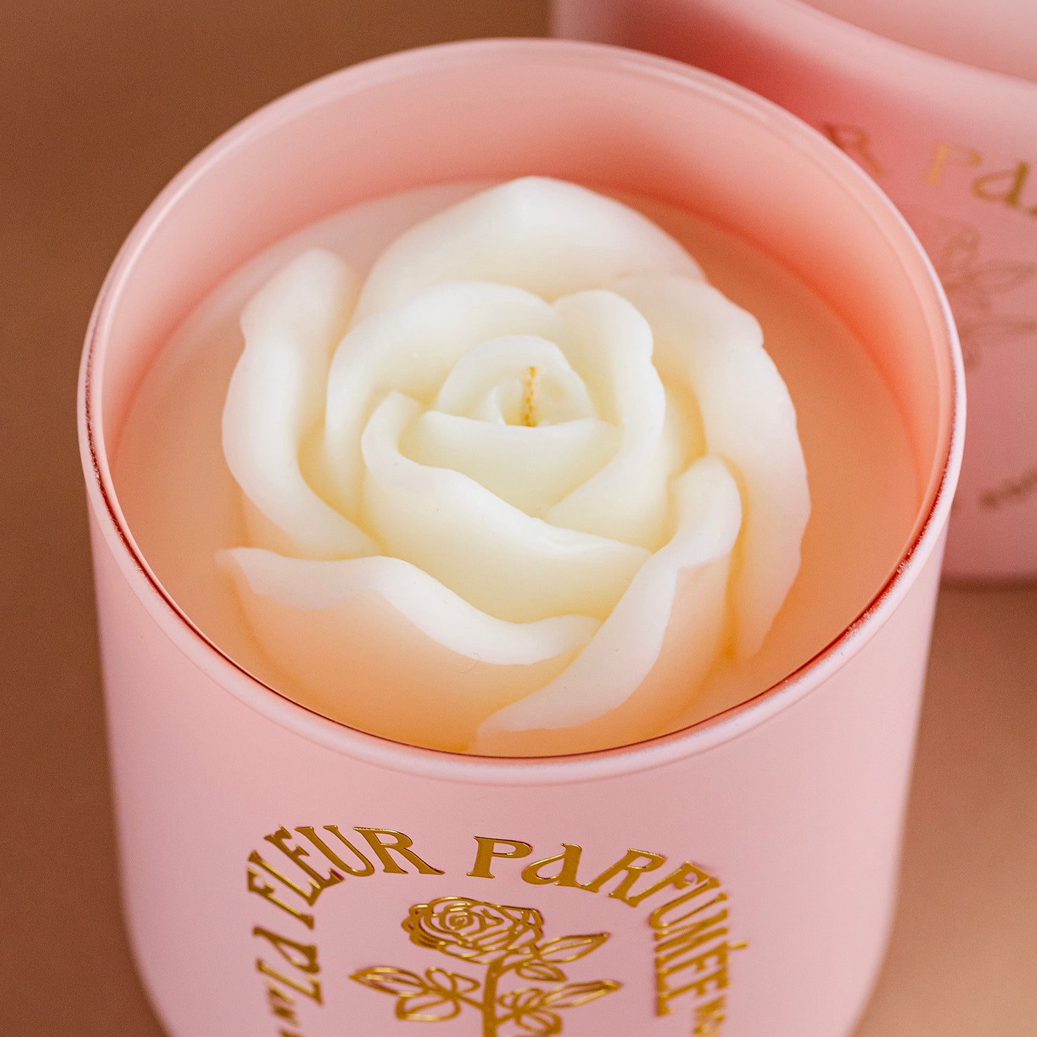 An elegant luxurious light pink candle with a gold emblem that reads La Fleur Parfumée An Expression of Adoration, and is topped with a white sculpted wax rose