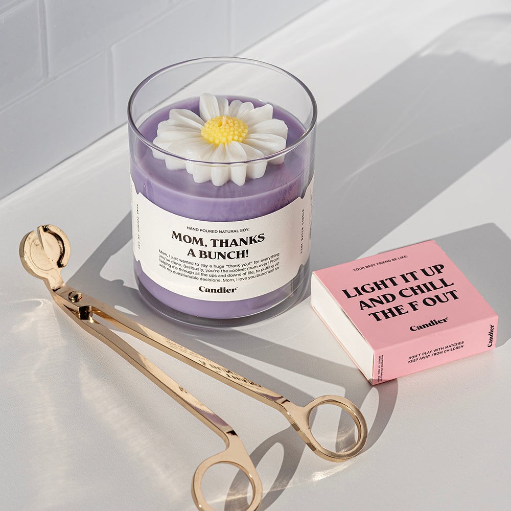 A purple candle with a sculpted wax flower on top and a label that reads Mom Thanks A Bunch. Beside it is a gold candle wick trimmer and a box of pink matches