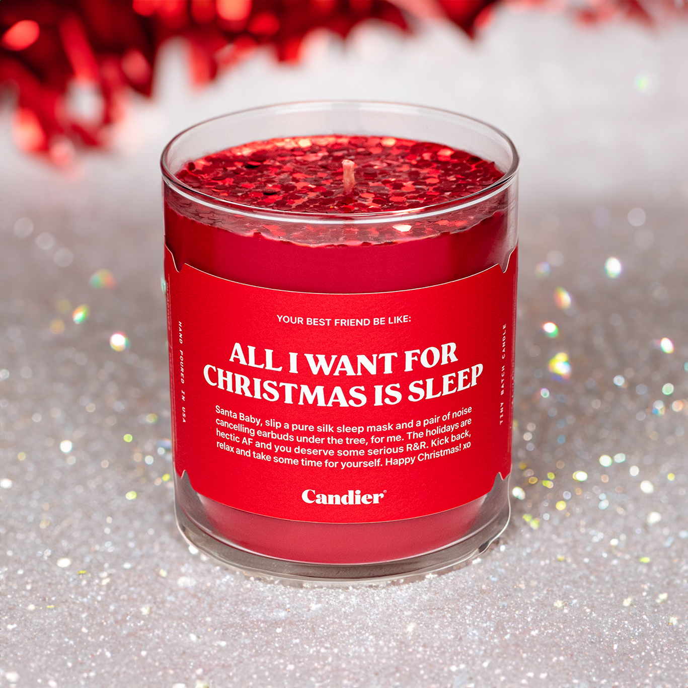 A Christmassy scene with a Candier candle in a clear glass vessel, filled with festive red wax, topped with sparkling red glitter, and a label that reads "All I Want For Christmas Is Sleep"