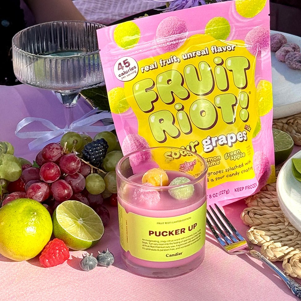Fruit Riot Pucker Up candle with citrus fruits and candy packaging in whimsical picnic setting. A best citrus scent candle from Candier candles, merging candycore and lux candle vibes.