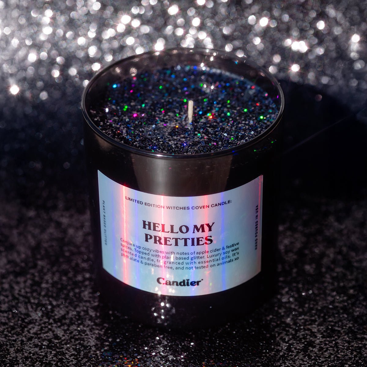 CandierĀ® Hello My Pretties candle on black glitter background, glowing with rich eco glitter wax, a luxe Halloween candle for wicked aesthetics and pretty candle lovers.