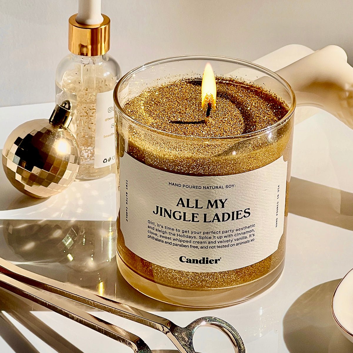A lit glitter gold Christmas themed scented candle with a label that reads ALL MY JINGLE LADIES by Candier