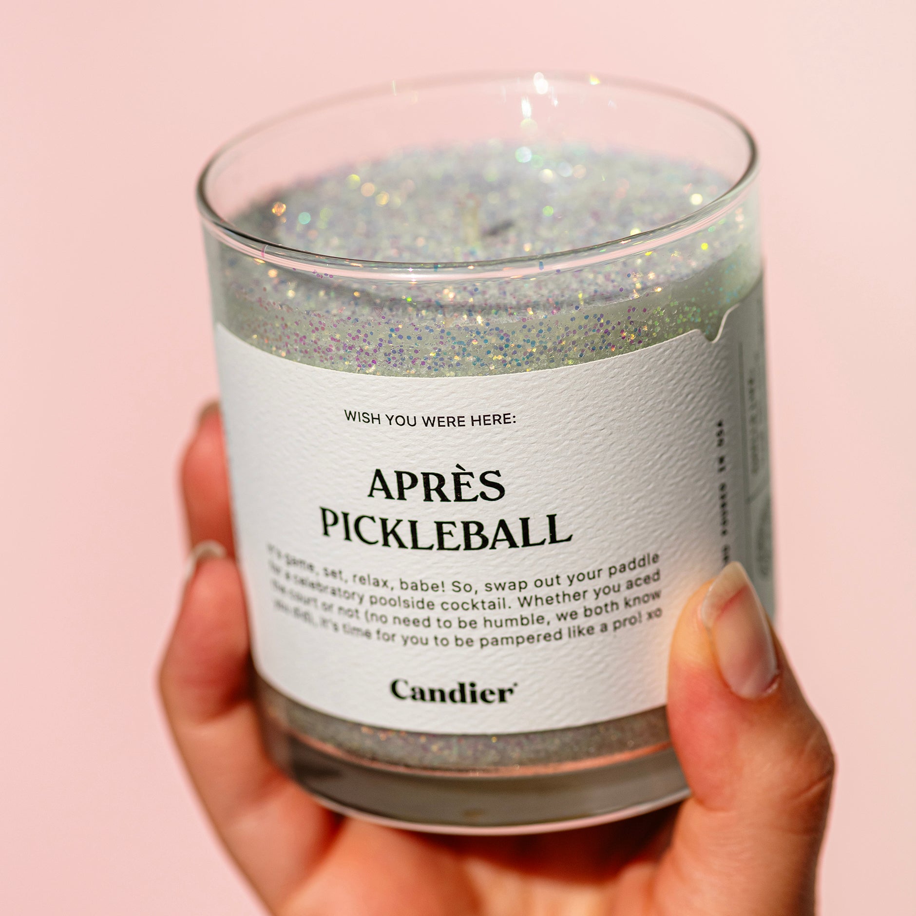 A hand holding a sparkling green candle with plant based glitter and a label that reads Aprés Pickleball. pickleball gifts for her​