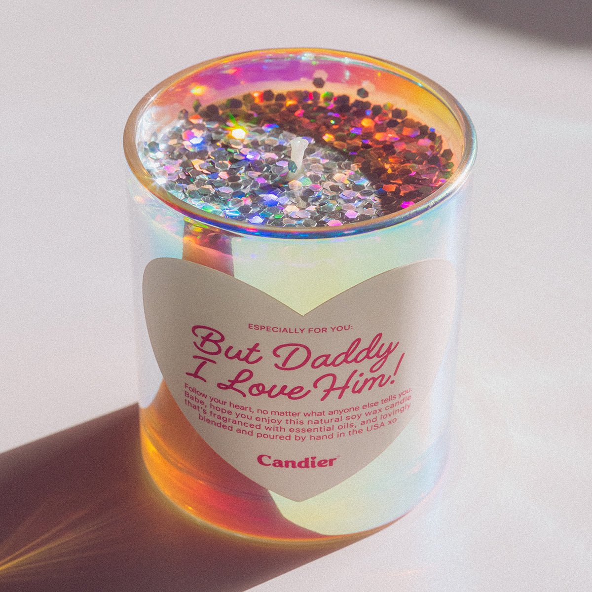 A shiny metallic scented candle with sparkling plant based glitter, and a heart shaped label that reads But Daddy, I Love Him by Candier®