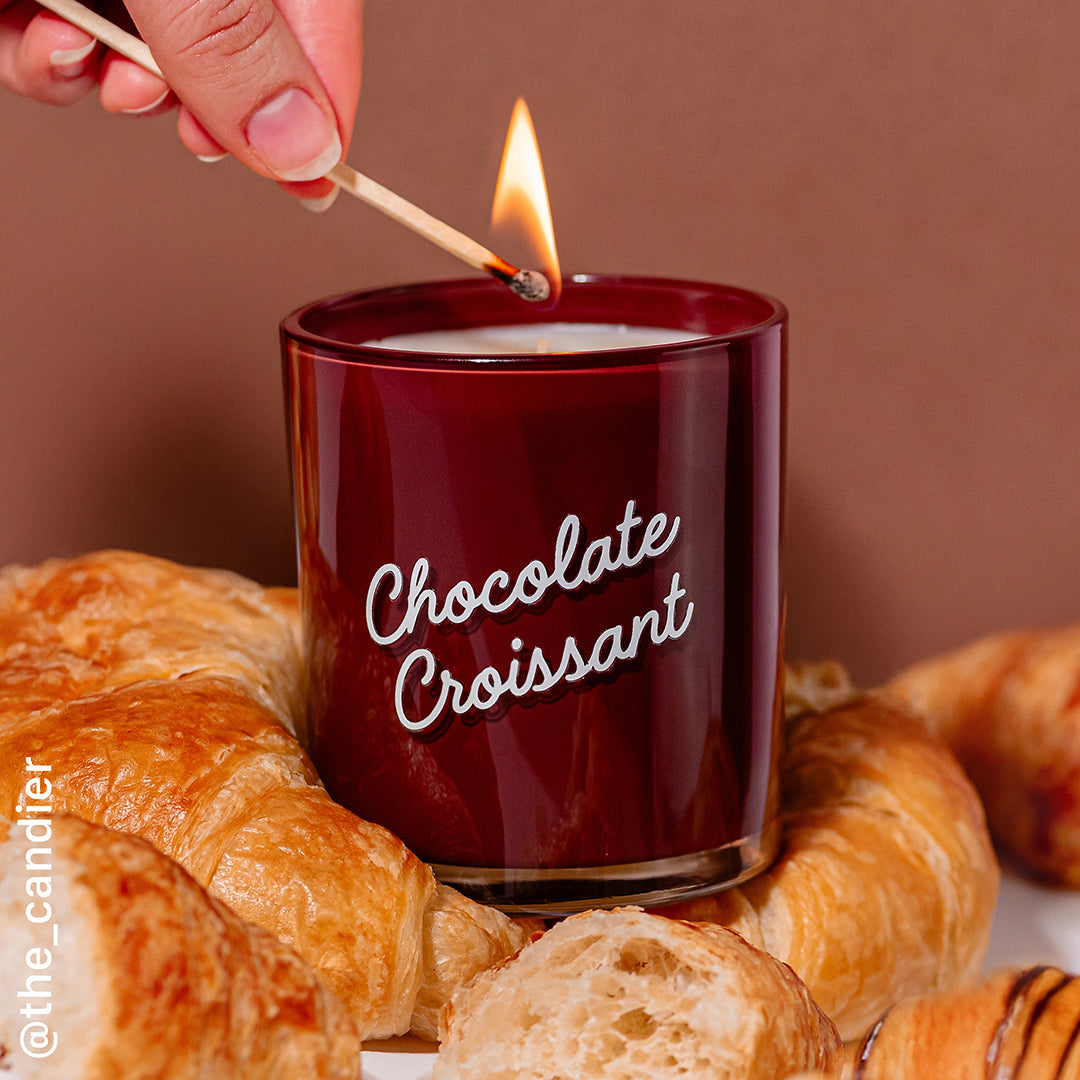 Hand lighting the Candier Chocolate Croissant candle next to fresh pastries. A cozy, chocolate candle with vanilla fragrance and hazelnut notes.