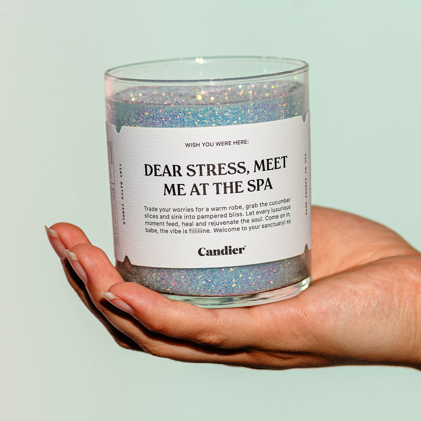 Hand holding a sparkling ocean blue candle with plant based glitter and a label that reads Dear Stress Met Me At The Spa
