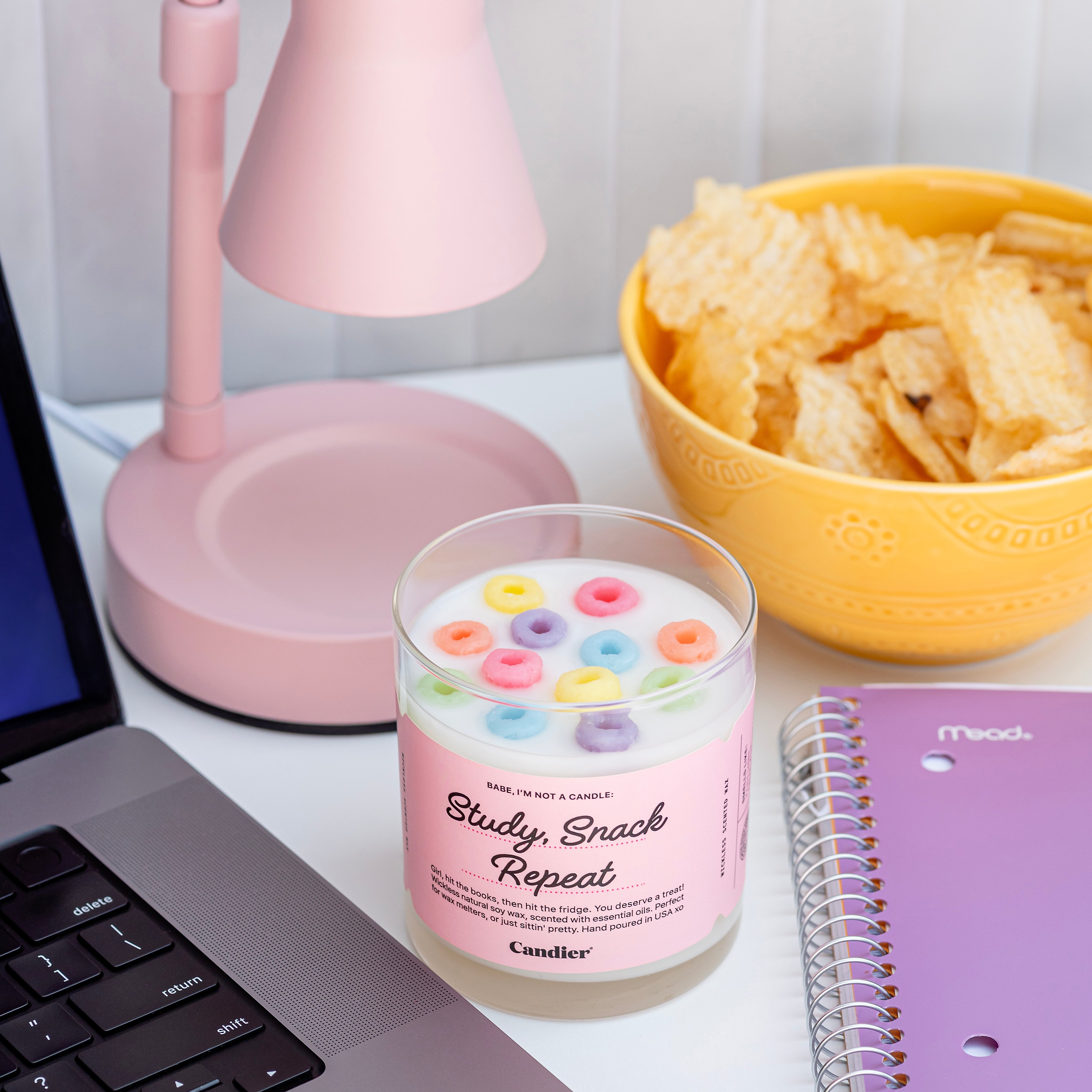 A wickless candle wax melt topped with colorful wax fruit loop cereal, and a label that reads Study, Snack, Repeat. Dormables by CANDIER, with a background of an Apple macbook , a pink wax melter lamp and a bowl of potato chips