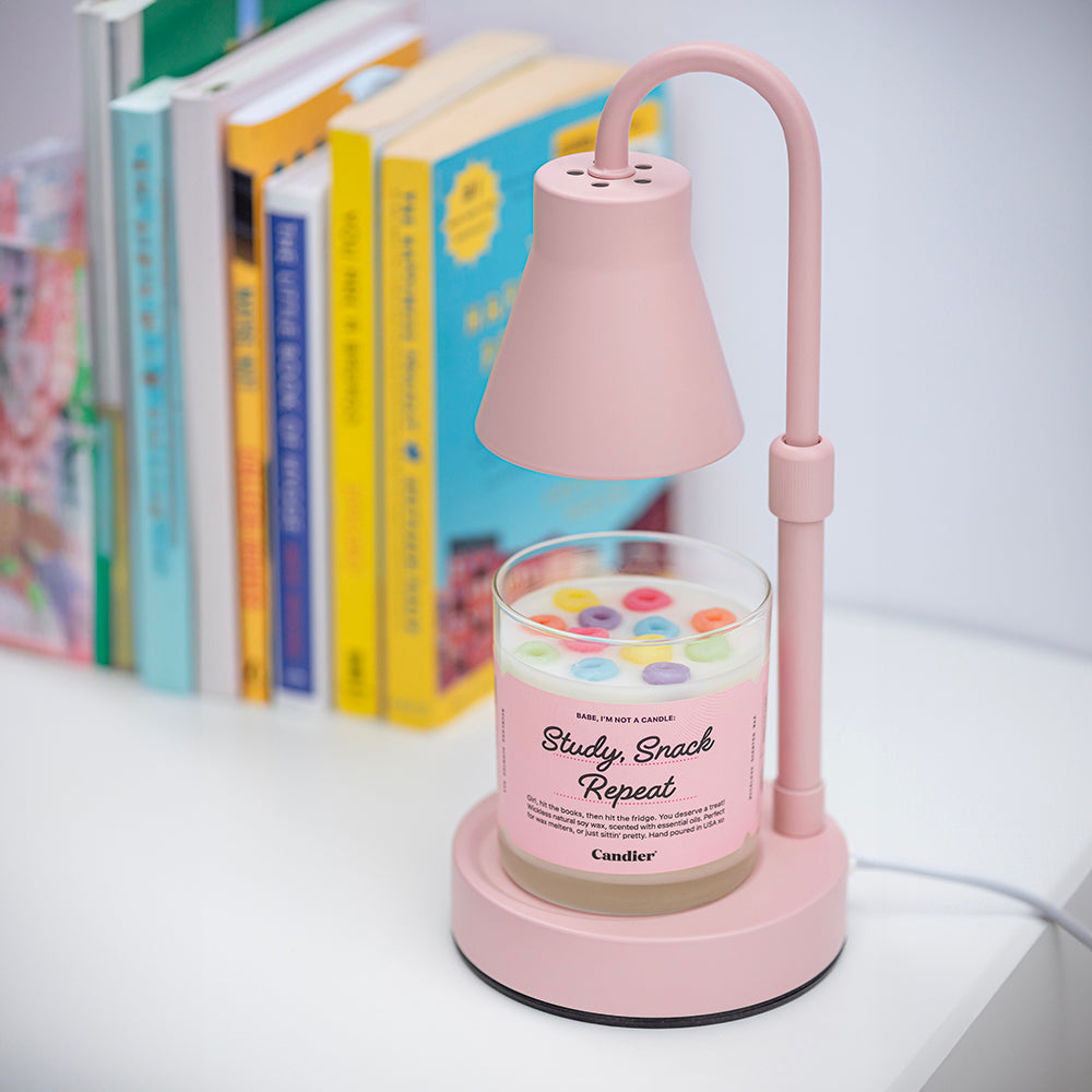 A wickless candle wax melt topped with colorful wax fruit loop cereal, and a label that reads Study, Snack, Repeat. Dormables by CANDIER, sitting on a pink wax melter lamp and a row of colorful books