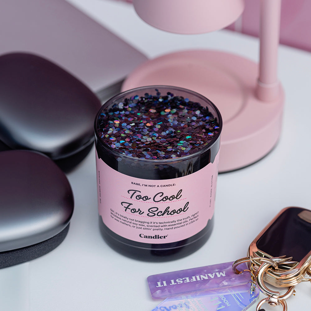 a glossy black wickless candle wax melt with a pink label that reads Too Cool For School. Dormables by CANDIER, sitting beside a pink lamp style wax melter