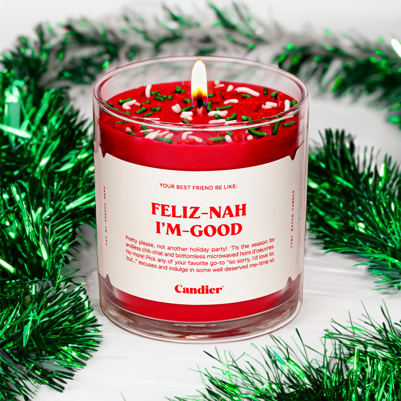 A lit Candier candle in a clear glass vessel, filled with Santa red wax, topped with red, green and white sprinkles, and a white label that reads “Feliz Nah I’m Good”. Green Christmas tinsel in the background