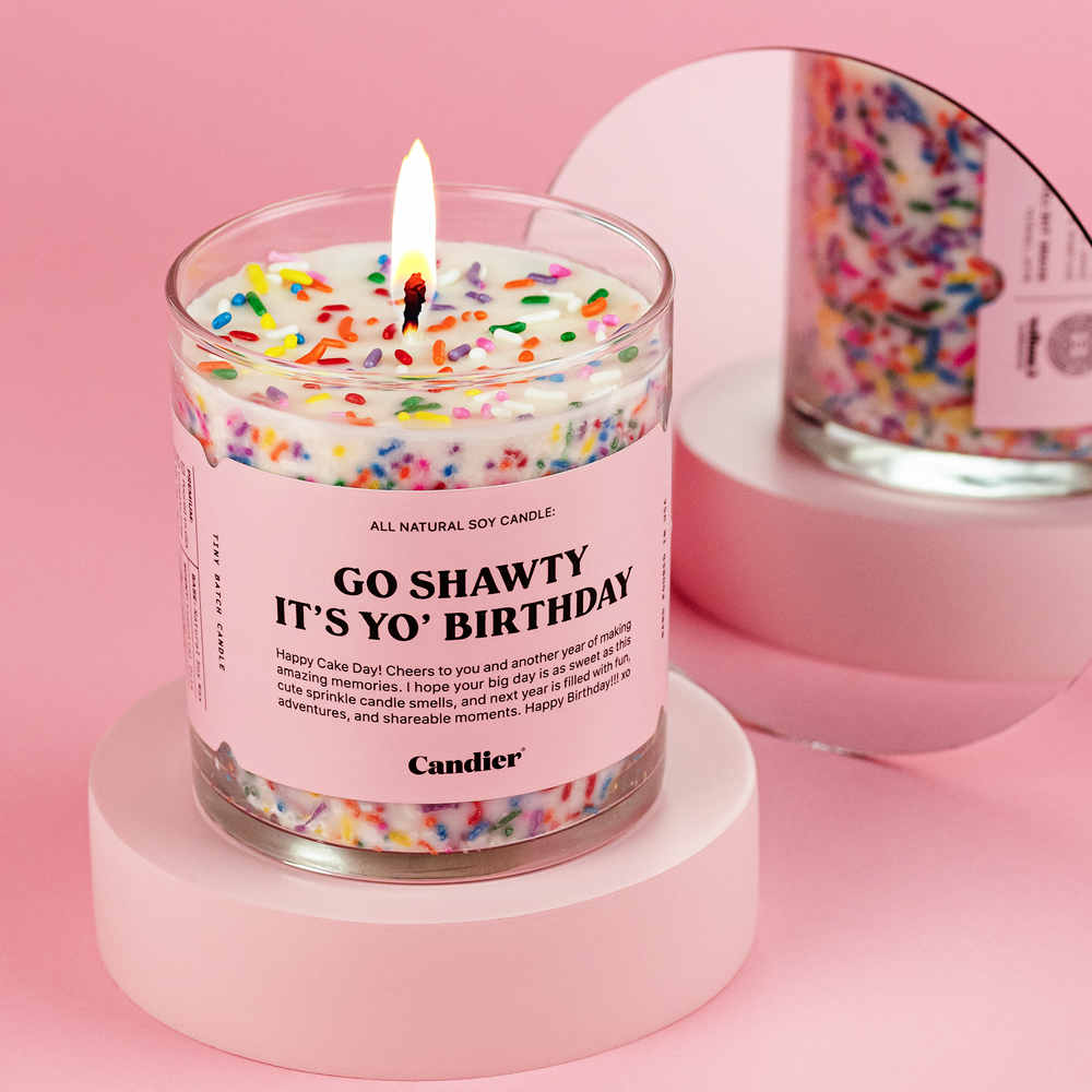 A luxury soy candle with a lit wick, labeled "GO SHAWTY IT'S YO BIRTHDAY," adorned with colorful sprinkles on top, resembling a festive birthday cake.