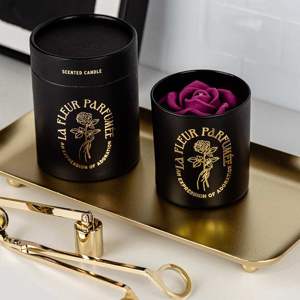 An fancy luxe black candle with a gold emblem that reads La Fleur Parfumée An Expression of Adoration, and is topped with a purple sculpted wax rose. It is surrounded by gold candle wick trimmer and snuffer and an elegant gift box