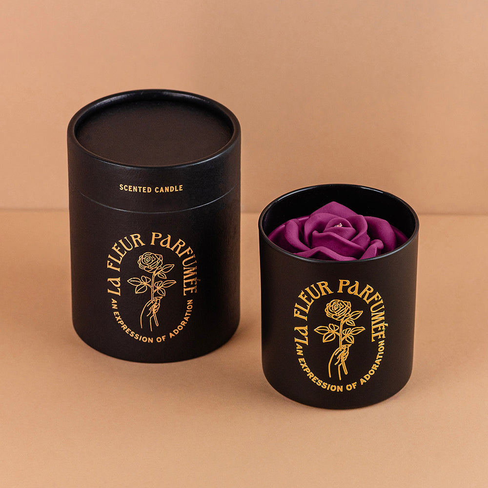 An fancy luxe black candle with a gold emblem that reads La Fleur Parfumée An Expression of Adoration, and is topped with a purple sculpted wax rose. It is beside an elegant gift box