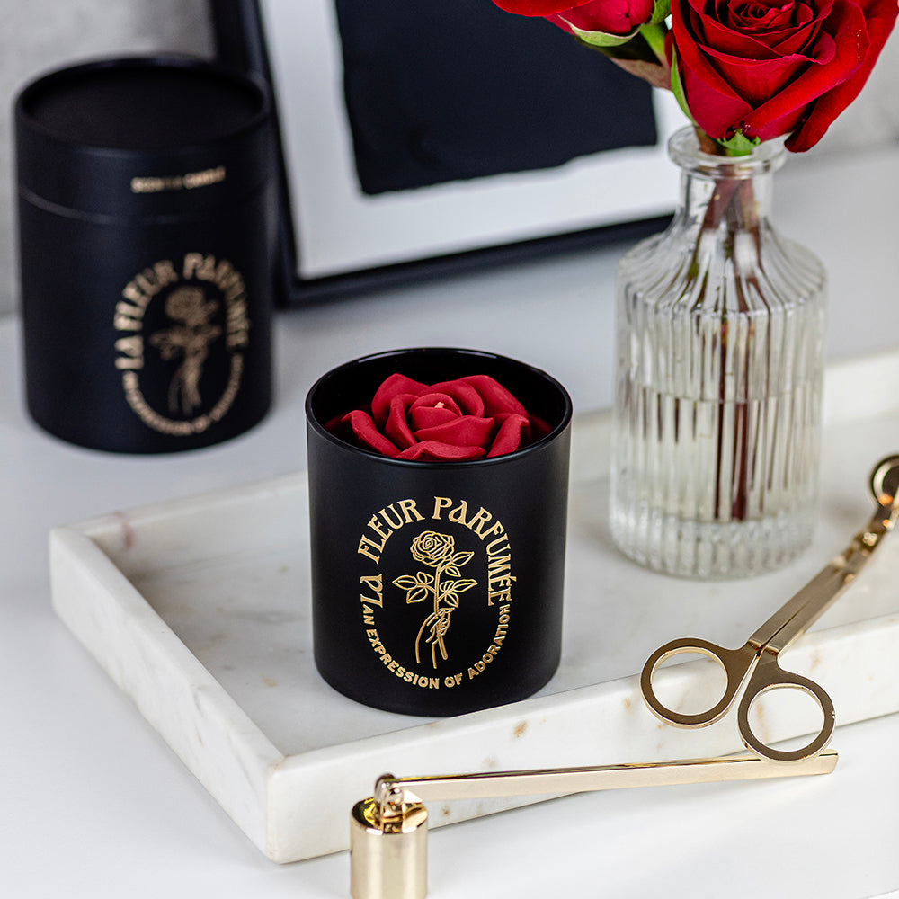 An fancy luxe black candle with a gold emblem that reads La Fleur Parfumée An Expression of Adoration, and is topped with a red sculpted wax rose. It is surrounded by gold candle wick trimmer and snuffer and an elegant gift box