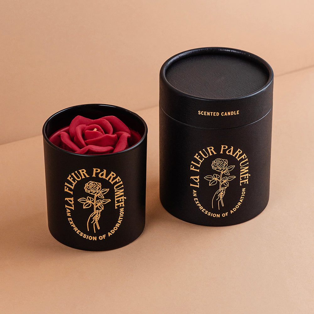 An fancy luxe black candle with a gold emblem that reads La Fleur Parfumée An Expression of Adoration, and is topped with a red sculpted wax rose. It is beside an elegant gift box
