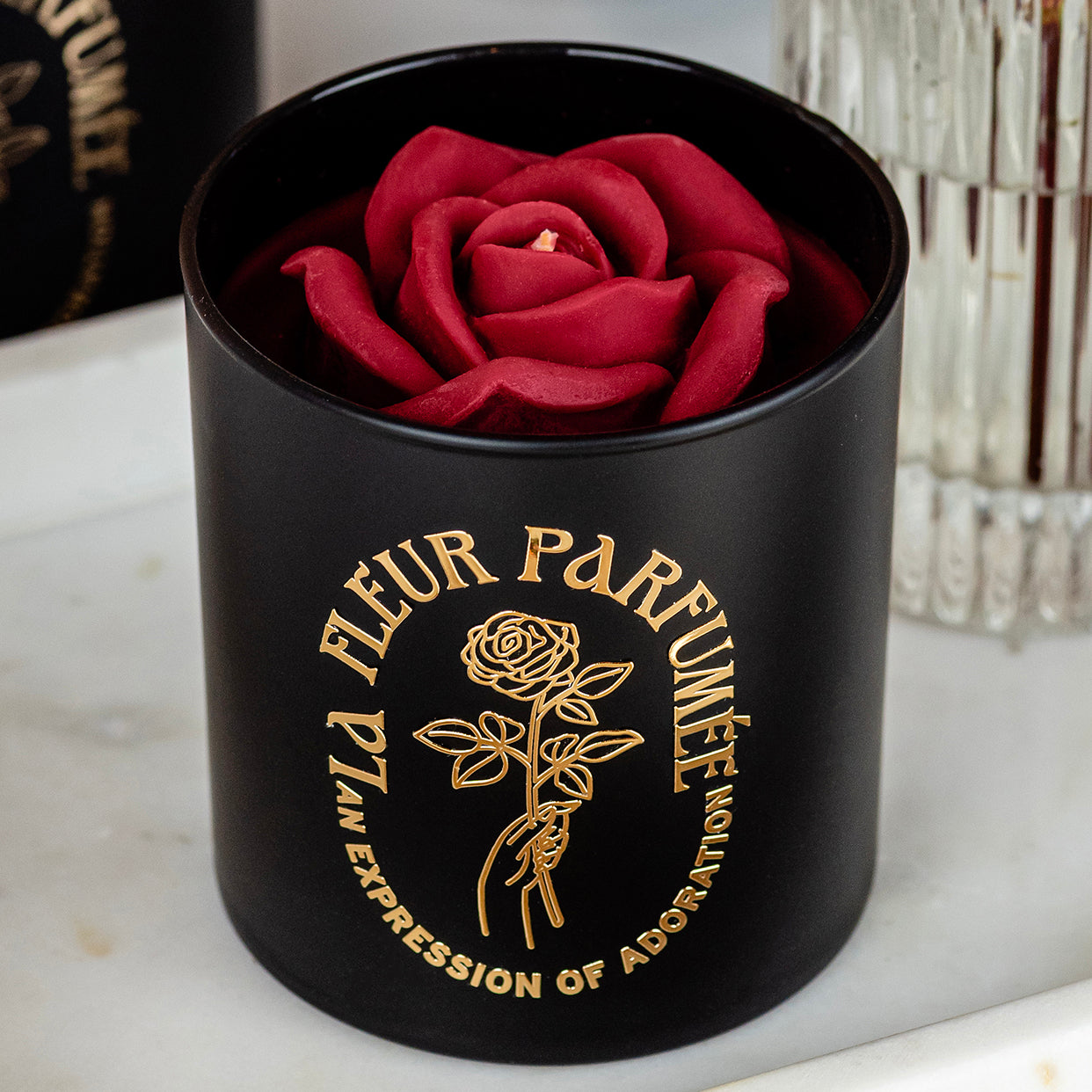 An elegant luxurious black candle with a gold emblem that reads La Fleur Parfumée An Expression of Adoration, and is topped with a red sculpted wax rose