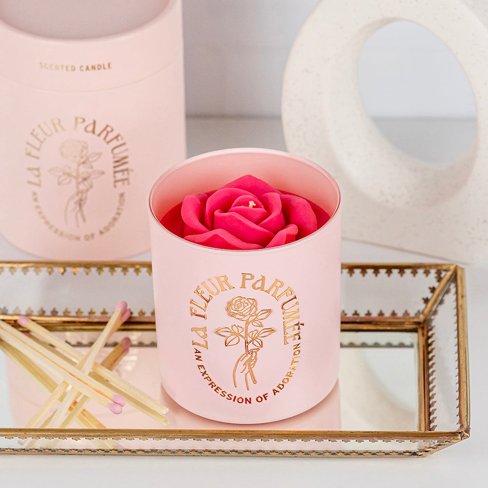 An elegant luxurious light pink candle with a gold emblem that reads La Fleur Parfumée An Expression of Adoration, and is topped with a hot pink sculpted wax rose. It sits on a glass tray and has pink tipped matches placed beside it