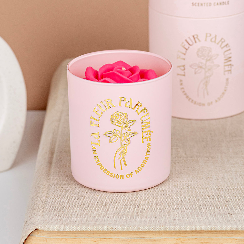 An elegant luxurious light pink candle with a gold emblem that reads La Fleur Parfumée An Expression of Adoration, and is topped with a hot pink sculpted wax rose