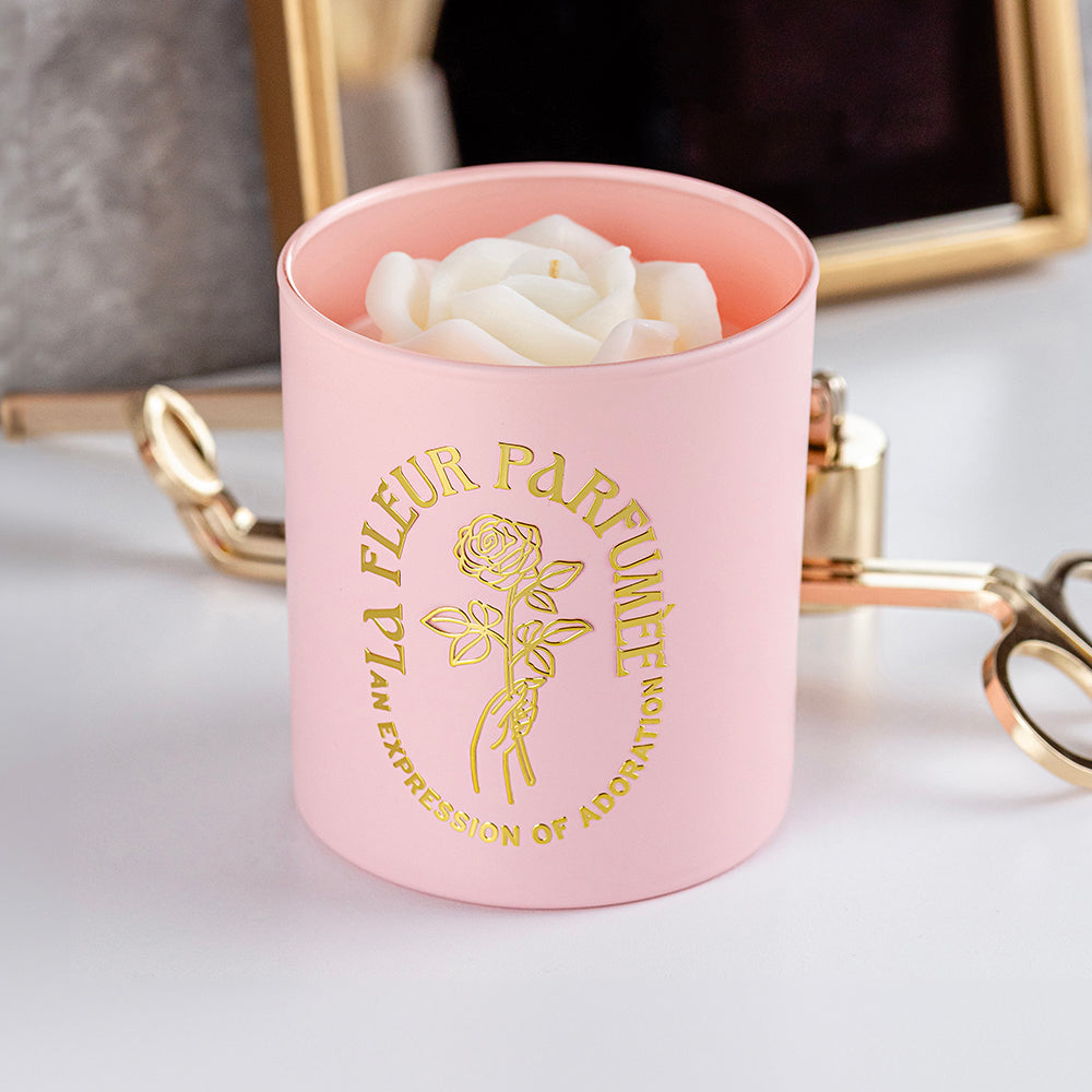 An elegant luxurious light pink candle with a gold emblem that reads La Fleur Parfumée An Expression of Adoration, and is topped with a white sculpted wax rose. Beside it is a gold wick trimmer and candle snuffer