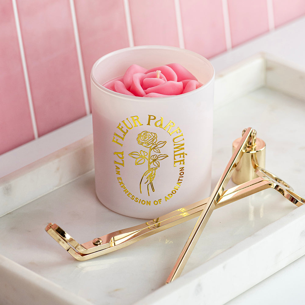 An elegant luxurious ivory white candle with a gold emblem that reads La Fleur Parfumée An Expression of Adoration, and is topped with a light blush pink sculpted wax rose. It sits on a white marble tray with a gold wick trimmer and candle snuffer