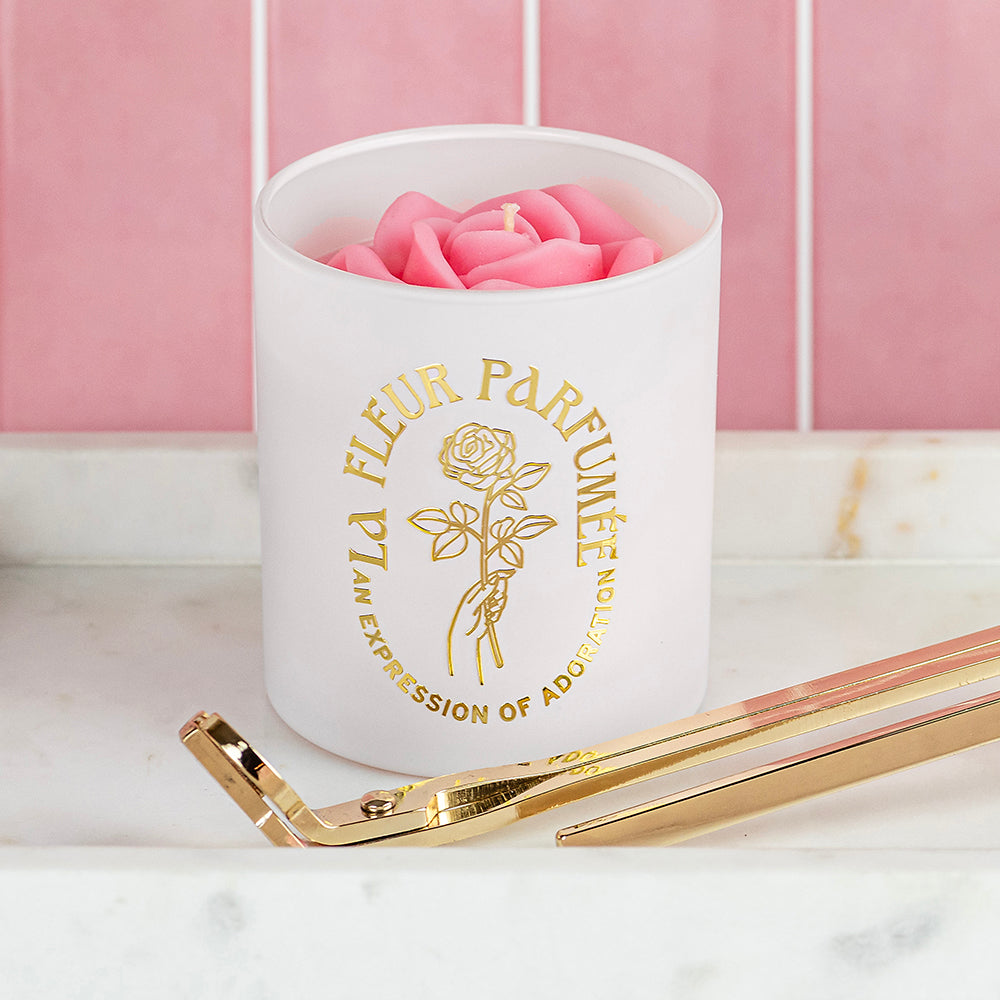 An elegant luxurious ivory white candle with a gold emblem that reads La Fleur Parfumée An Expression of Adoration, and is topped with a light blush pink sculpted wax rose. It sits on a white marble tray with a gold wick trimmer and candle snuffer