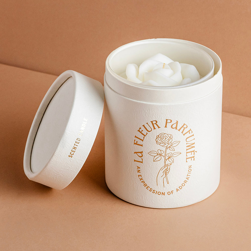 An elegant luxurious ivory white candle with a gold emblem that reads La Fleur Parfumée An Expression of Adoration, and is topped with an ivory white sculpted wax rose. The candle sits inside a premium high end gift box
