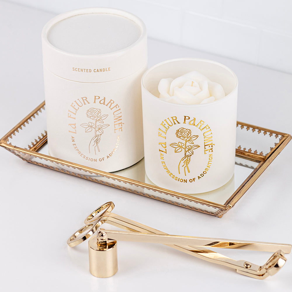 An elegant luxurious ivory white candle with a gold emblem that reads La Fleur Parfumée An Expression of Adoration, and is topped with an ivory white sculpted wax rose. It sits on a glass try, beside a gift box and candle trimmer and snuffer