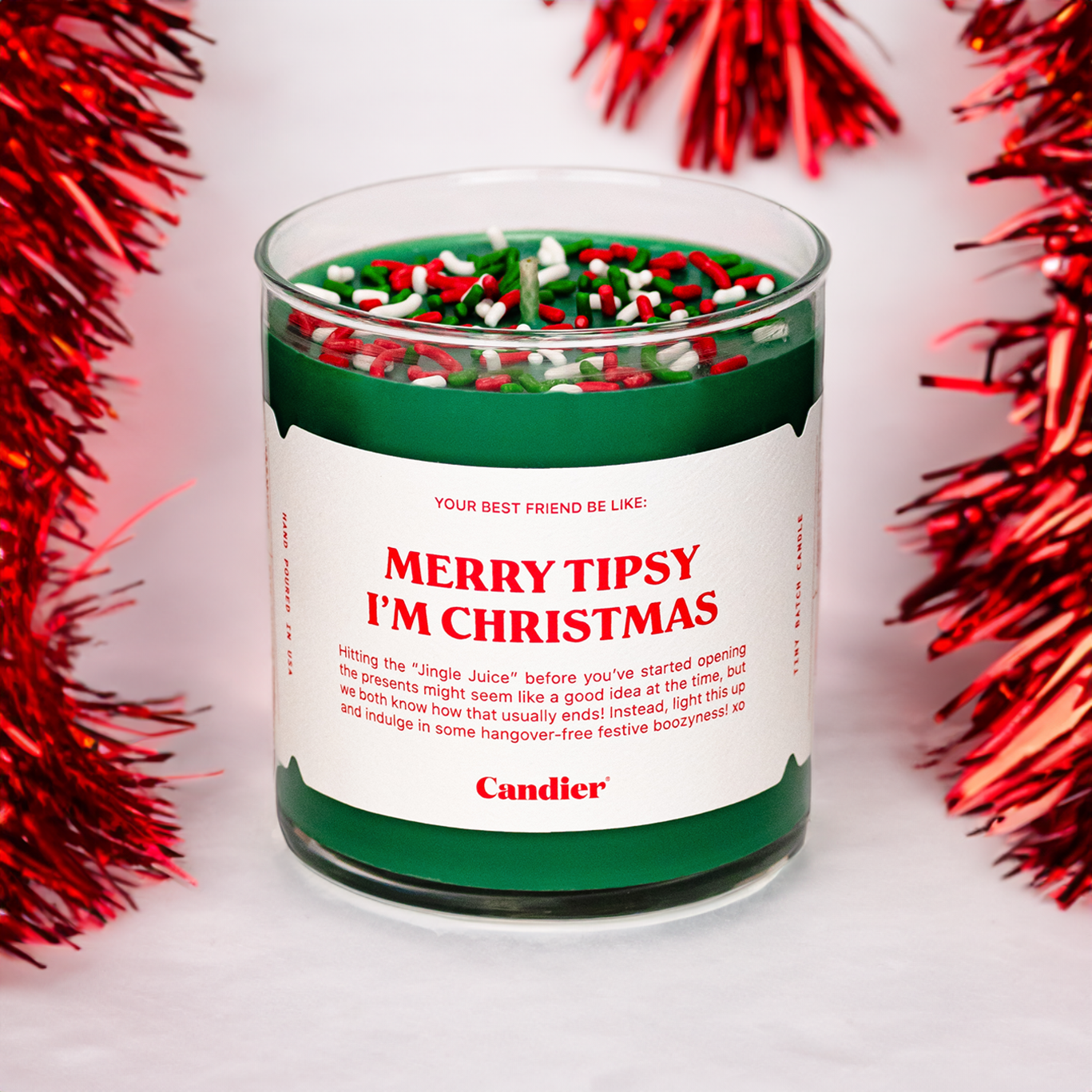 A Christmas themed Candier candle in a clear glass vessel, filled with festive green wax, topped with red, green and white sprinkles, and a white label that reads “Merry tipsy, I’m Christmas”. Red tinsel in background
