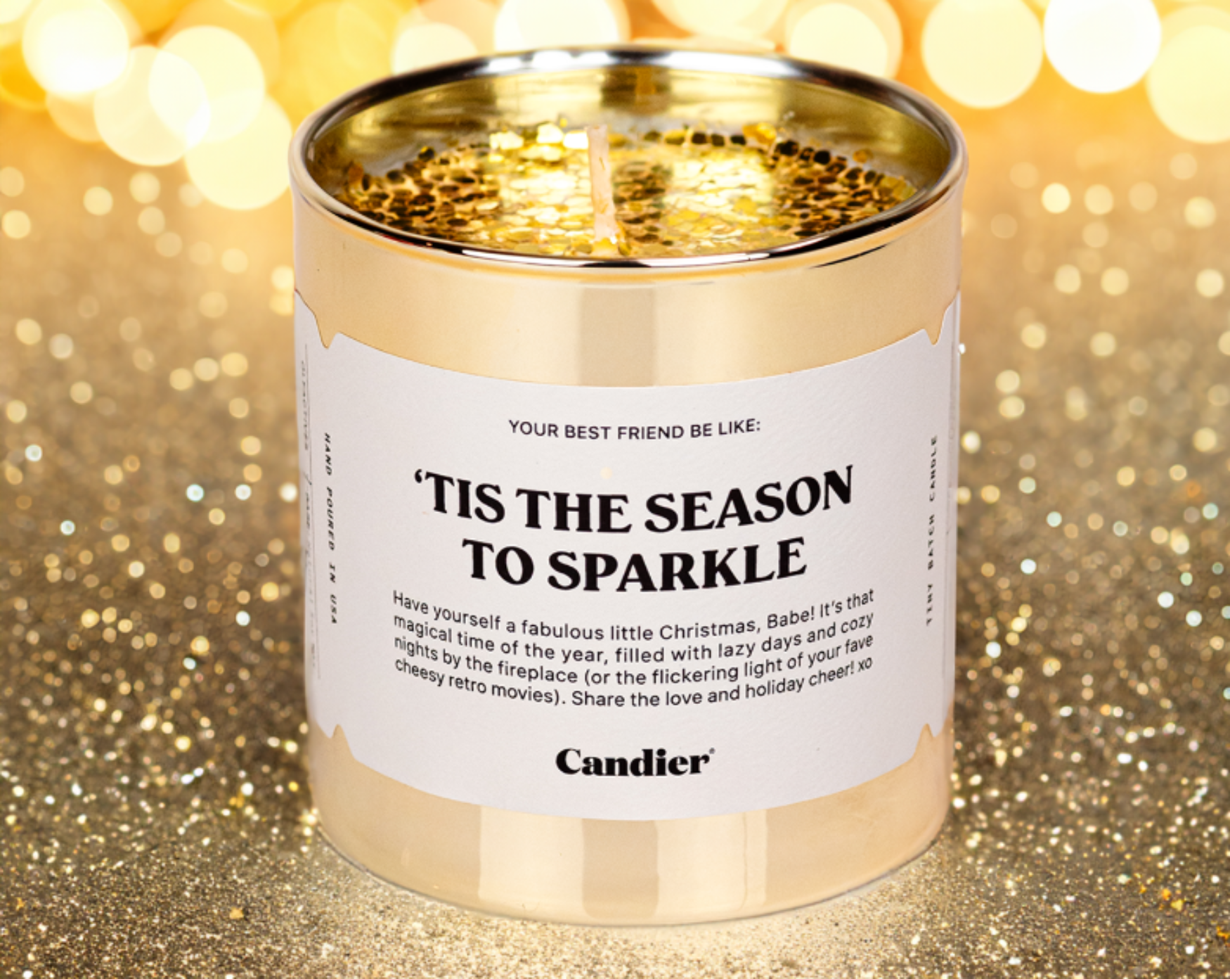 A Christmas themed Candier candle in a metallic gold glass vessel, topped with and surrounded by sparkling gold glitter and a label that reads "Tis The Season To Sparkle"